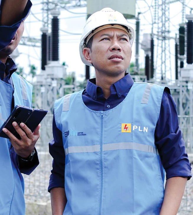 Two men wearing blue vests are standing in front of a power plant. One man is pointing at a cell phone, possibly showing the other man something important. AI generated content